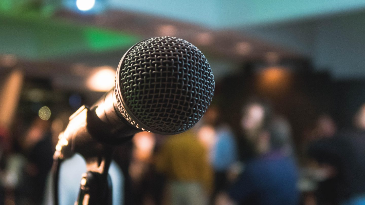 Close up shot of a microphone on a stand with blurred attendees in the background.