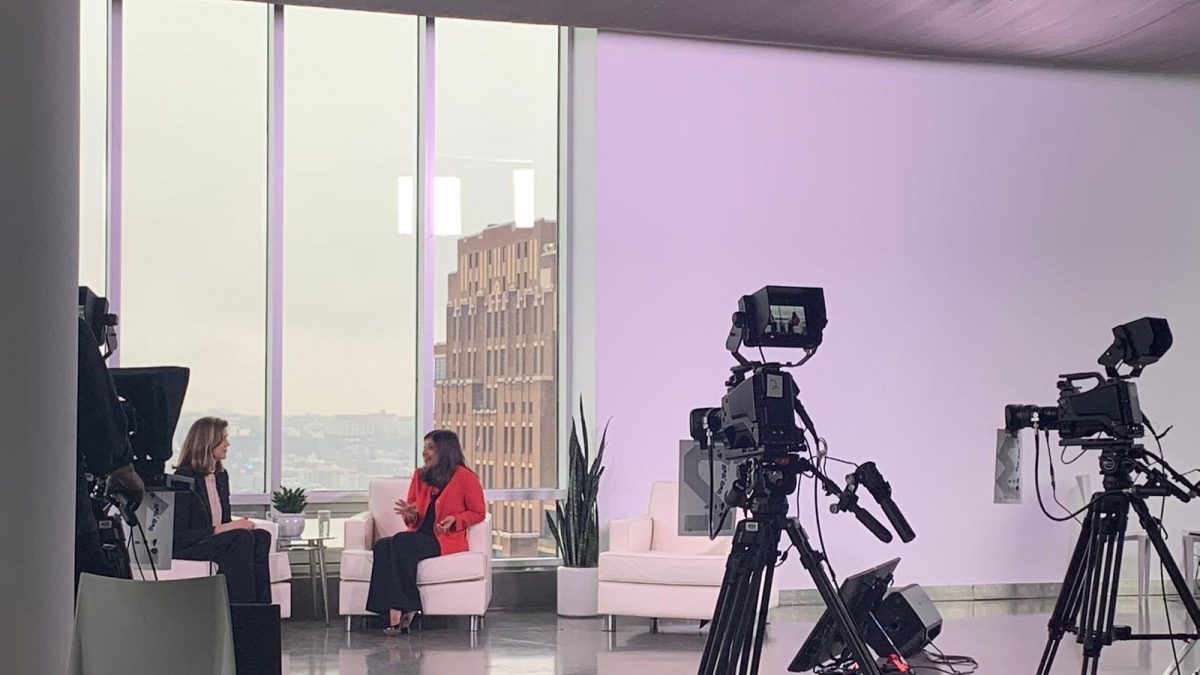 Cameras in a studio recording two women speaking to each other infront of a window showing buildings in a city