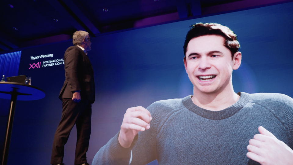 Man in suit  is on a stage looking up at a screen with a large avatar that is a man with brown hair